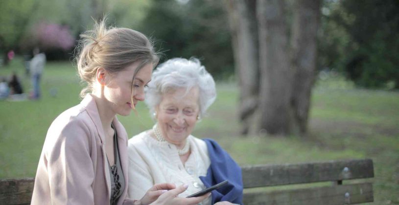 Senior woman with a younger female on a park bench, looking at her phone.