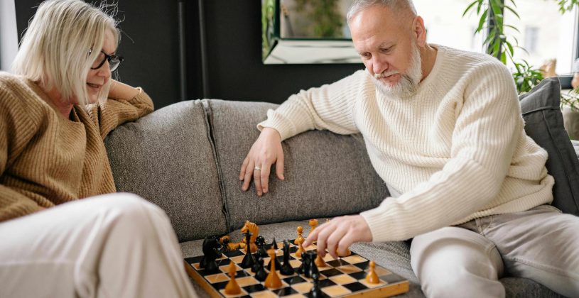 A senior couple playing a game of chess on a gray couch.